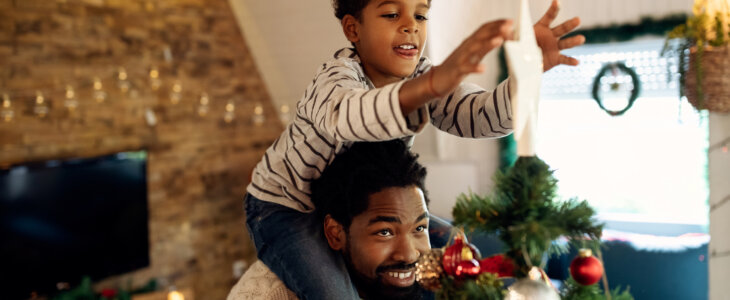 Happy African American father carrying his son on shoulders and helping him to put star on top of Christmas tree.